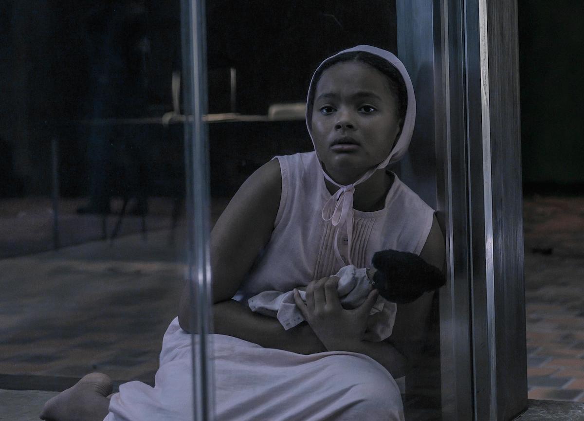 Jordana Blake in a pink dress and bonnet holding a doll while sitting in a glass room as Hannah in 'The Handmaid's Tale' Season 4