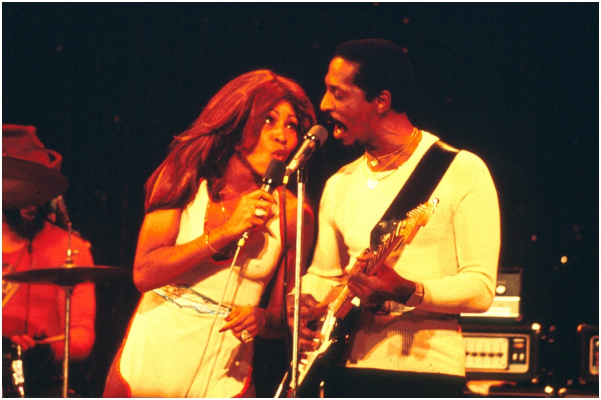 Tina Turner dancing and singing next to Ike Turner while wearing a white outfit. Ike is wearing a white shirt and black pants.