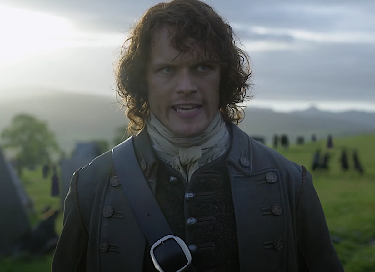 Sam Heughan in a 1700s coat and vest, a white scarf around his neck and a black strap across his chest as Jamie Fraser in 'Outlander.' He stands on a green field in front of tents on a sunny morning.