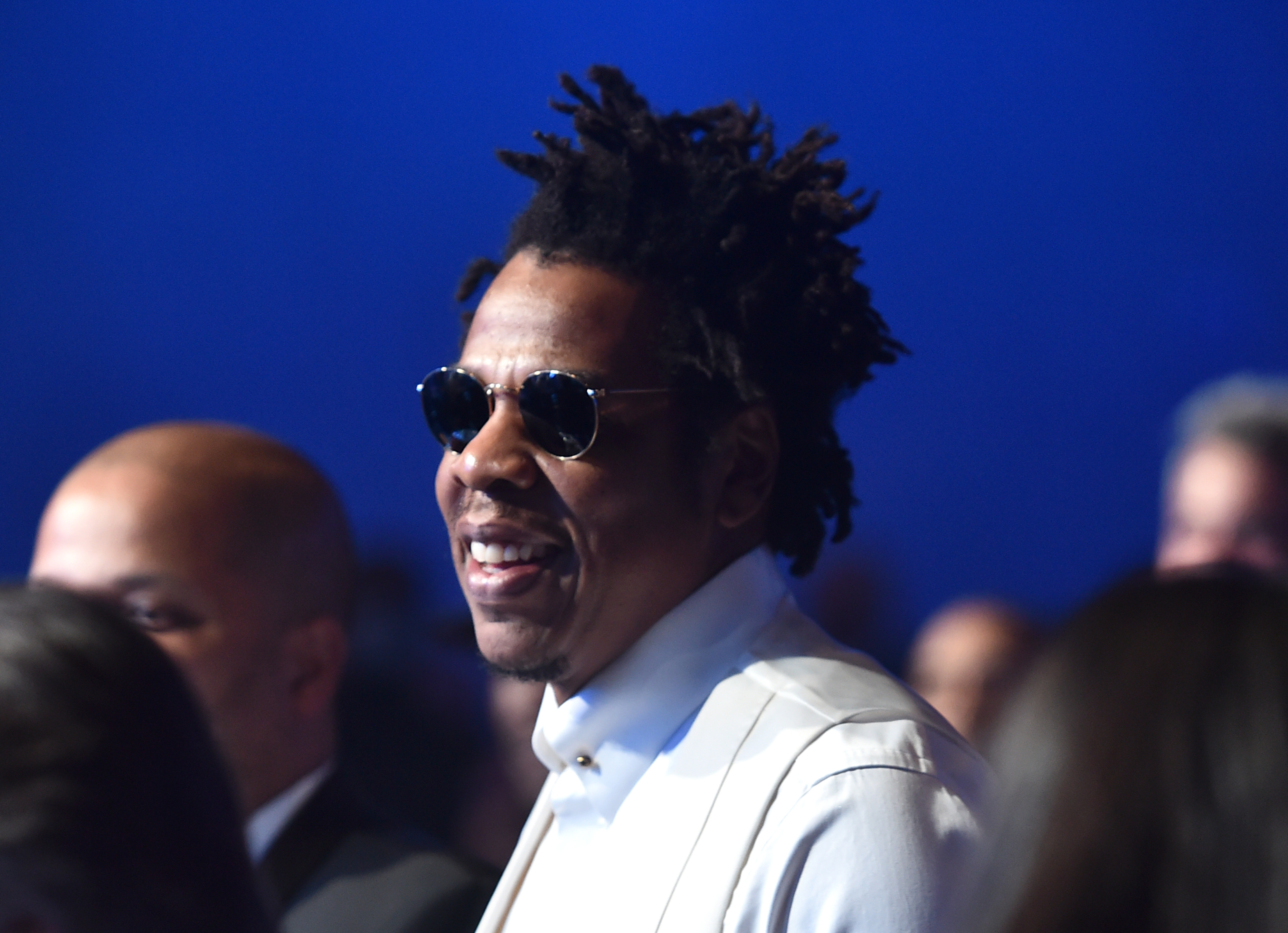 Jay-Z smiling while wearing black sunglasses and a white jacket at the 2020 Grammy Awards pre-show.