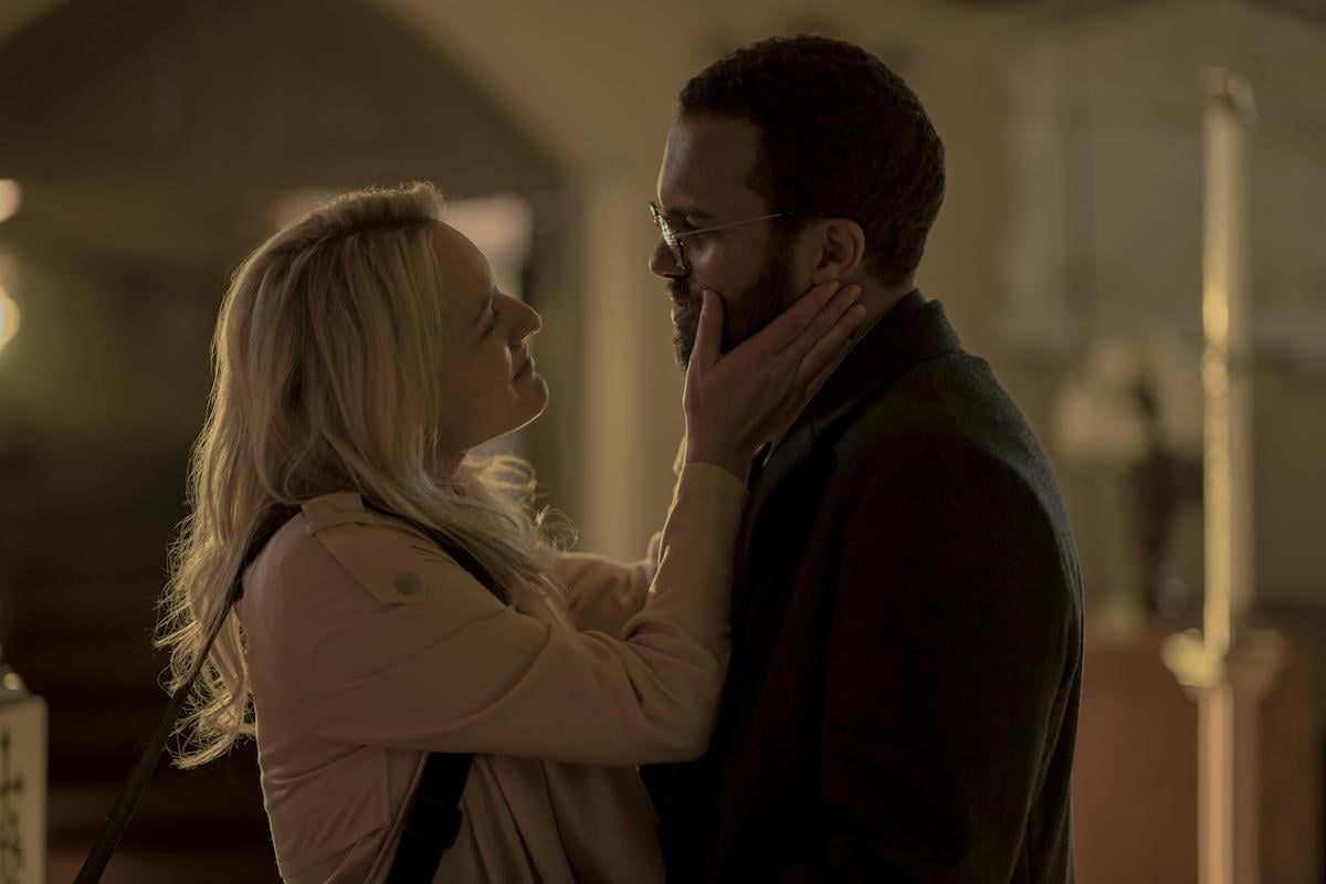 June (Elisabeth Moss) smiles and holds Luke's (O.T. Fagbenle) face between her hands in 'The Handmaid's Tale' Season 3. She wears a pink jacket and has a purse hanging over her shoulder and he wears a dark blazer and glasses.