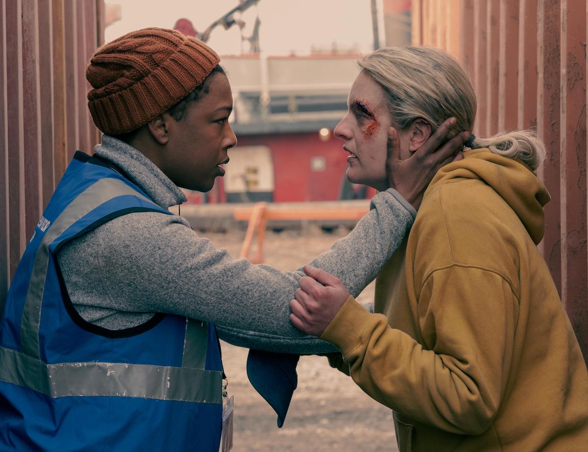 Samira Wiley as Moira in a blue vest, orange beanie, and light grey zip-up turtleneck sweater. She holds Elisabeth Moss face in her hands, who plays June. Moss wears a yellow hoodie and has dirt and blood on her face. From season 4 of 'The Handmaid's Tale'