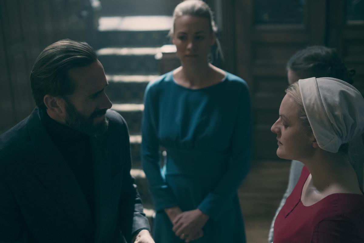 Commander Waterford (Joseph Fiennes) in a black turtleneck and grey suit, Serena Joy (Yvonne Strahovski) in a teal dress, and June Osborne (Elisabeth Moss) in a red dress and white bonnet in 'The Handmaid's Tale'