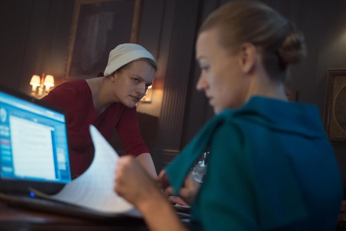 Elisabeth Moss in a red dress and white bonnet as June and Yvonne Strahovski in a teal dress as Serena Joy in 'The Handmaid's Tale' Season 2. Serena Joy sits at a desk as June watches her.