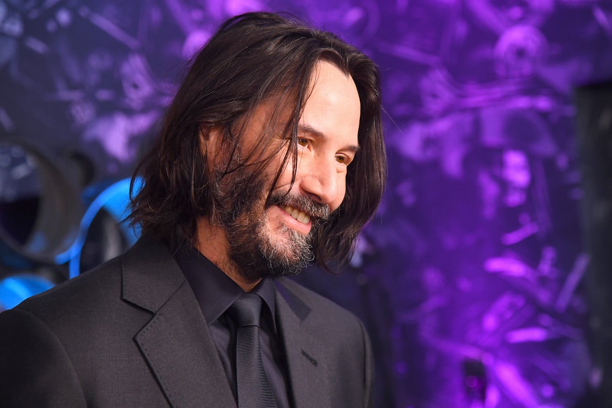 Keanu Reeves smiles in a black suit in front of a purple backdrop at 'John Wick' special screenings
