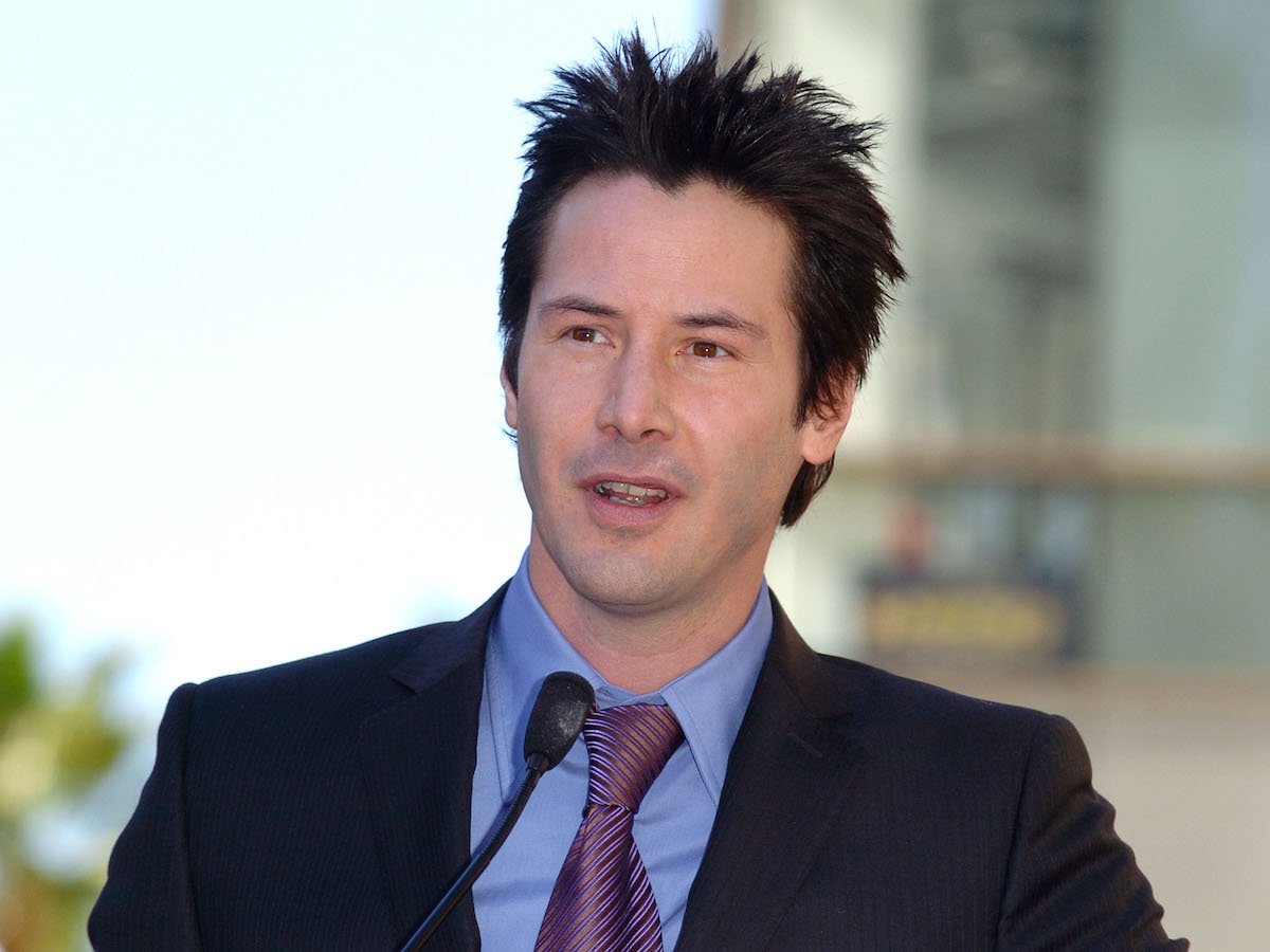 Keanu Reeves wears a suit and tie while speaking at a microphone