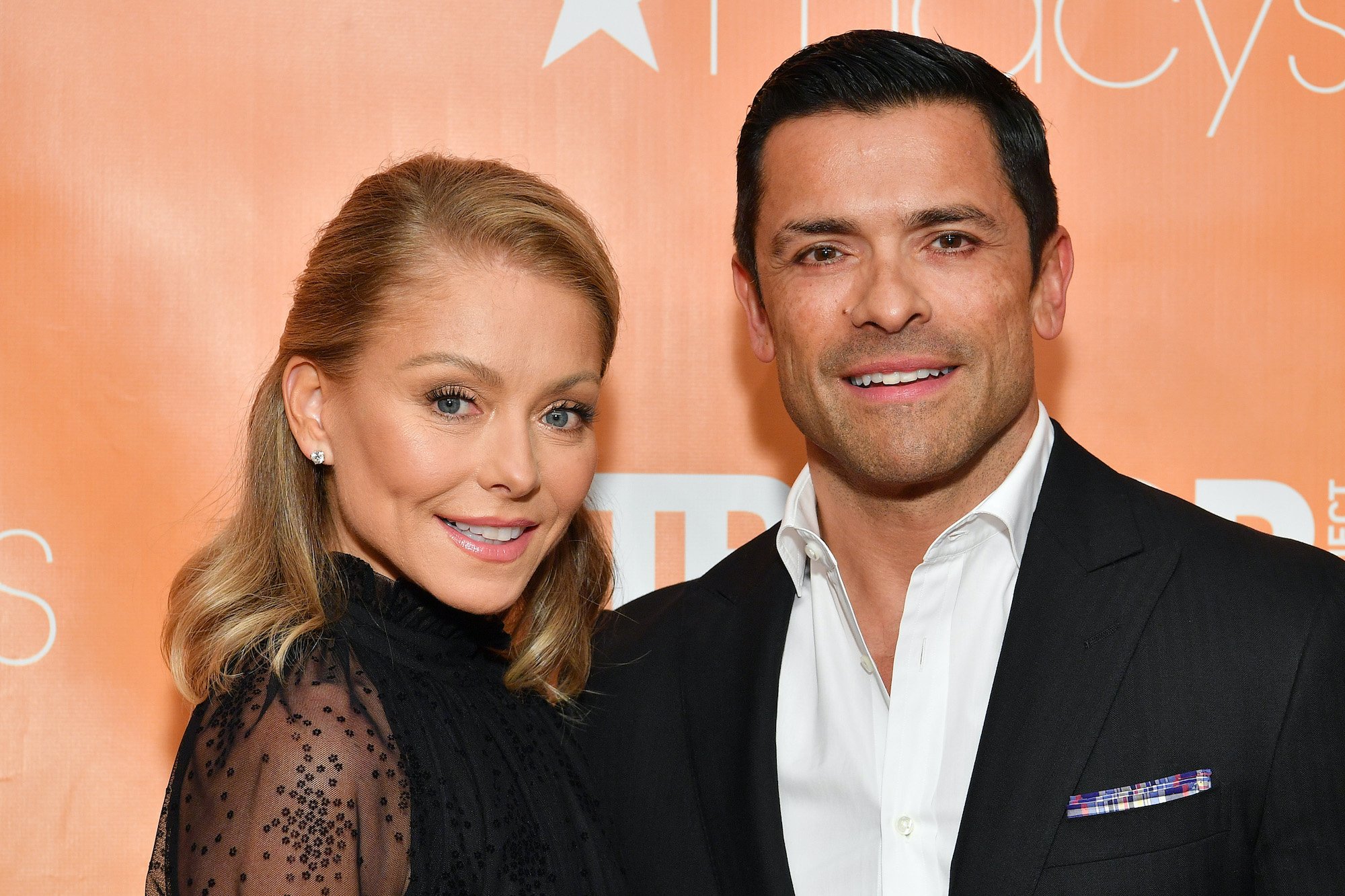 (L-R) Kelly Ripa and Mark Consuelos smiling in front of an orange background
