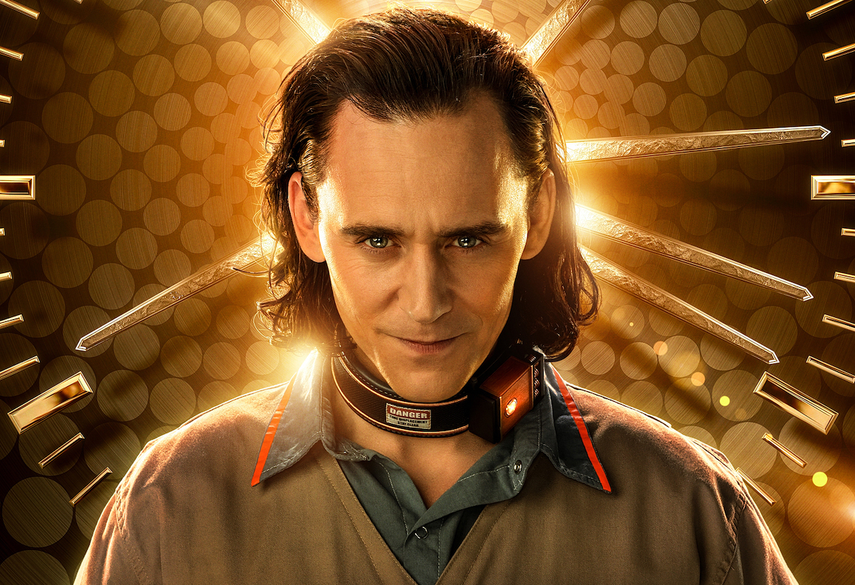 Tom Hiddleston as Loki in a beige and blue prison uniform with a monitor around his neck in front of a gold, clock-like background