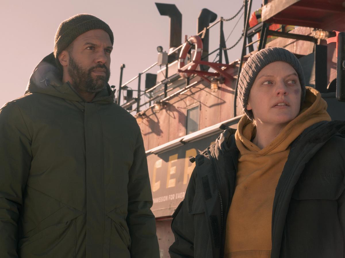 O-T Fagbenle as Luke wearing a green winter coat and beanie, and Elisabeth Moss as June wearing a yellow hoodie, green winter coat, and grey beanie as they stand in front of a boat in 'The Handmaid's Tale' Season 4.