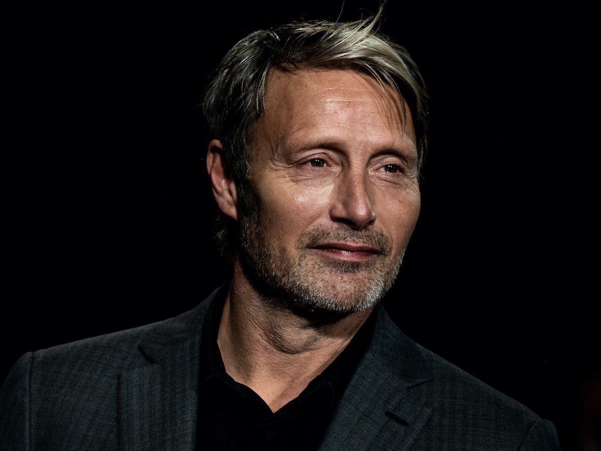 Mads Mikkelsen in a dark grey suit and black shirt in front of a black background