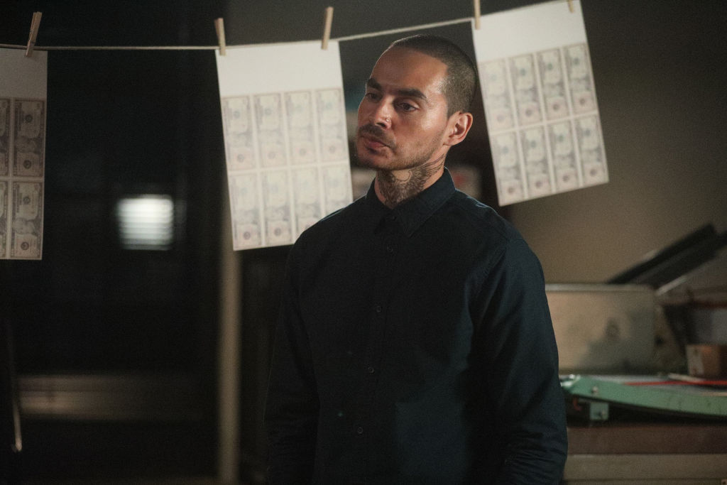 Manny Montana, who's dressed in all black, plays Rio on 'Good Girls'. He's standing in front of laundered money with a scowl.