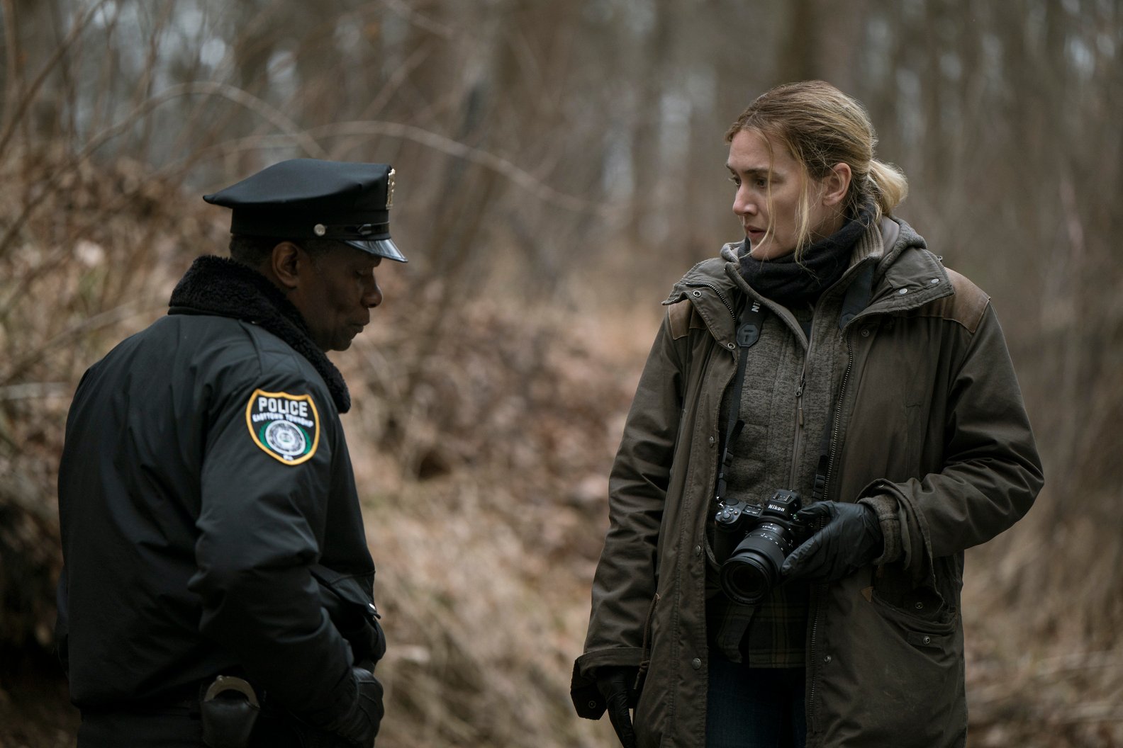 Kate Winslet plays small-town detective Mare who is talking to John Douglas Carter who plays Chief Carter as they investigate a murder in the woods on a dreary day in Easttown.