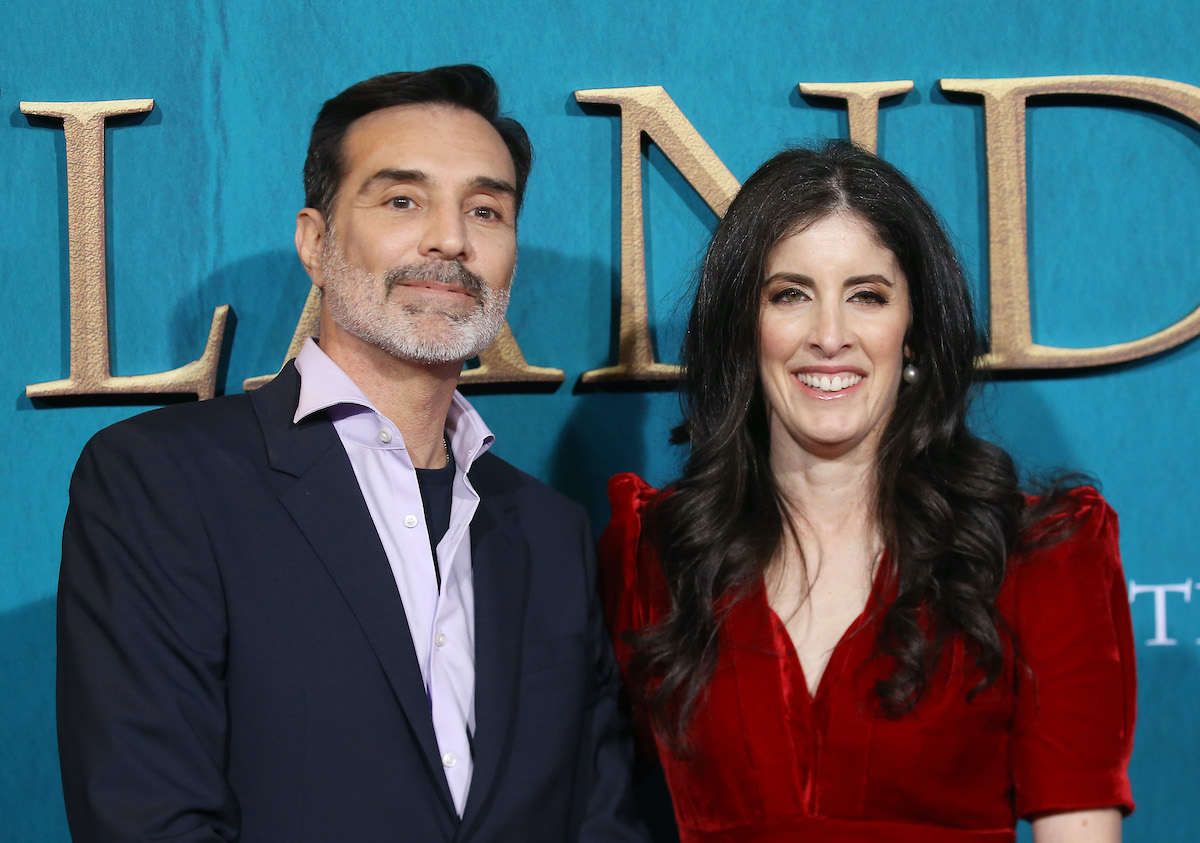 Matthew B. Roberts (L) in a lavender button-down shirt and navy blue blazer standing next to Maril Davis (R) in a red velvet dress with short sleeves and accented shoulders. Roberts and Davis are two of 'Outlander's executive producers. They stand in front of a cerulean backdrop that says 'Outlander' written in gold.