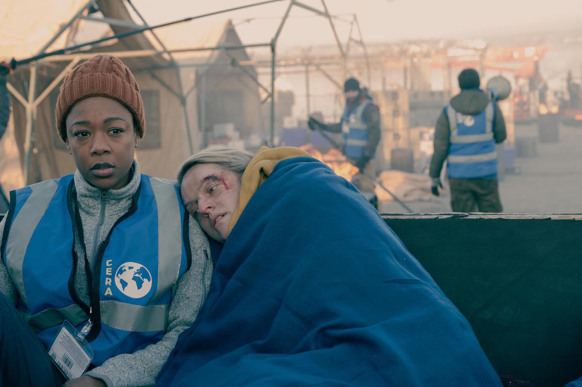 Samira Wiley as Moira in a blue vest, orange beanie, and light grey zip-up turtleneck sweater. Elisabeth Moss as June lays her head on Wiley's shoulder wearing a yellow hoodie and wrapped in a blue blanket. From season 4 of 'The Handmaid's Tale.'