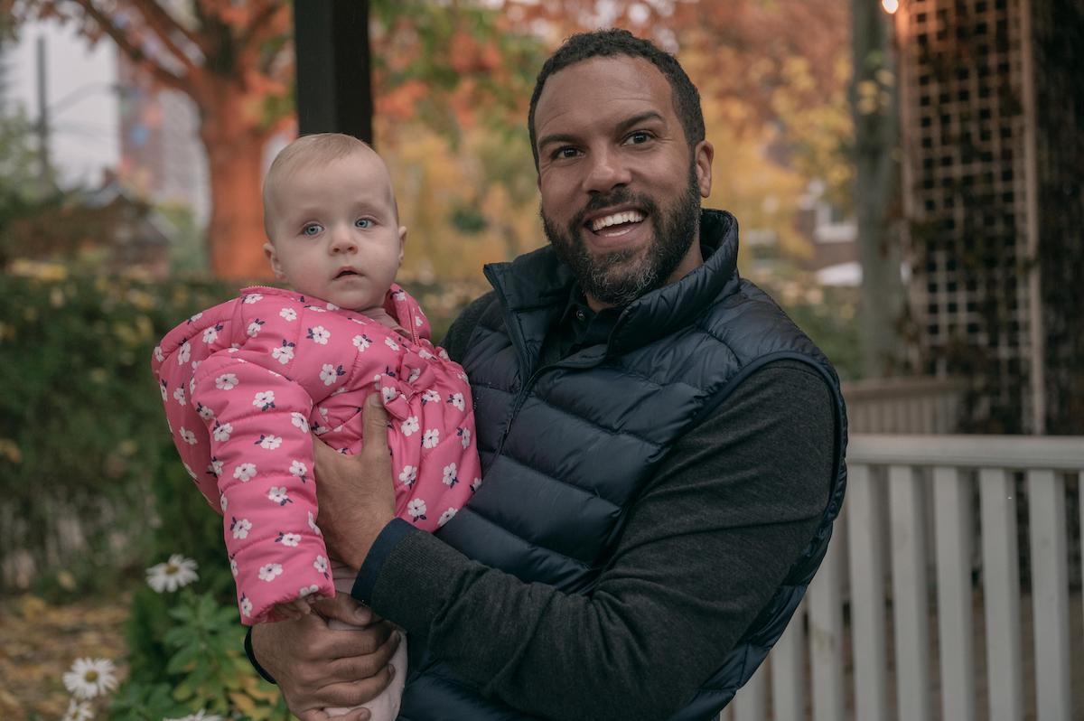 O-T Fagbenle as Luke in 'The Handmaid's Tale' holding baby Nicole. He's smiling and wearing a black shirt and puffer vest. Nicole wears a pink jacket.