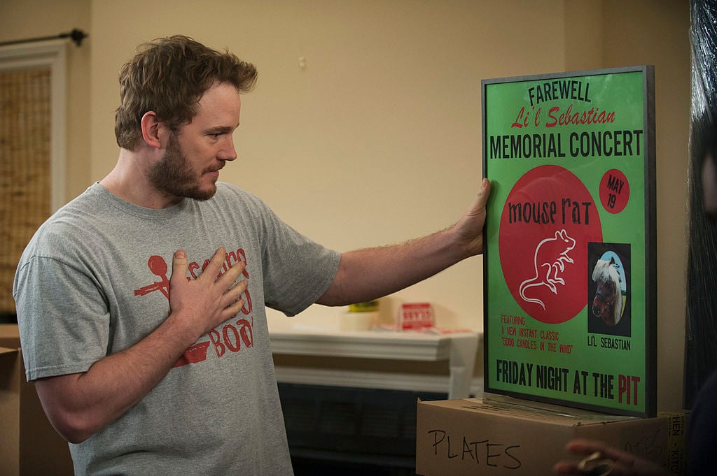 Chris Pratt as Andy Dwyer, dressed in a grey t-shirt, looks at a green and red poster for a Mouse Rat concert.