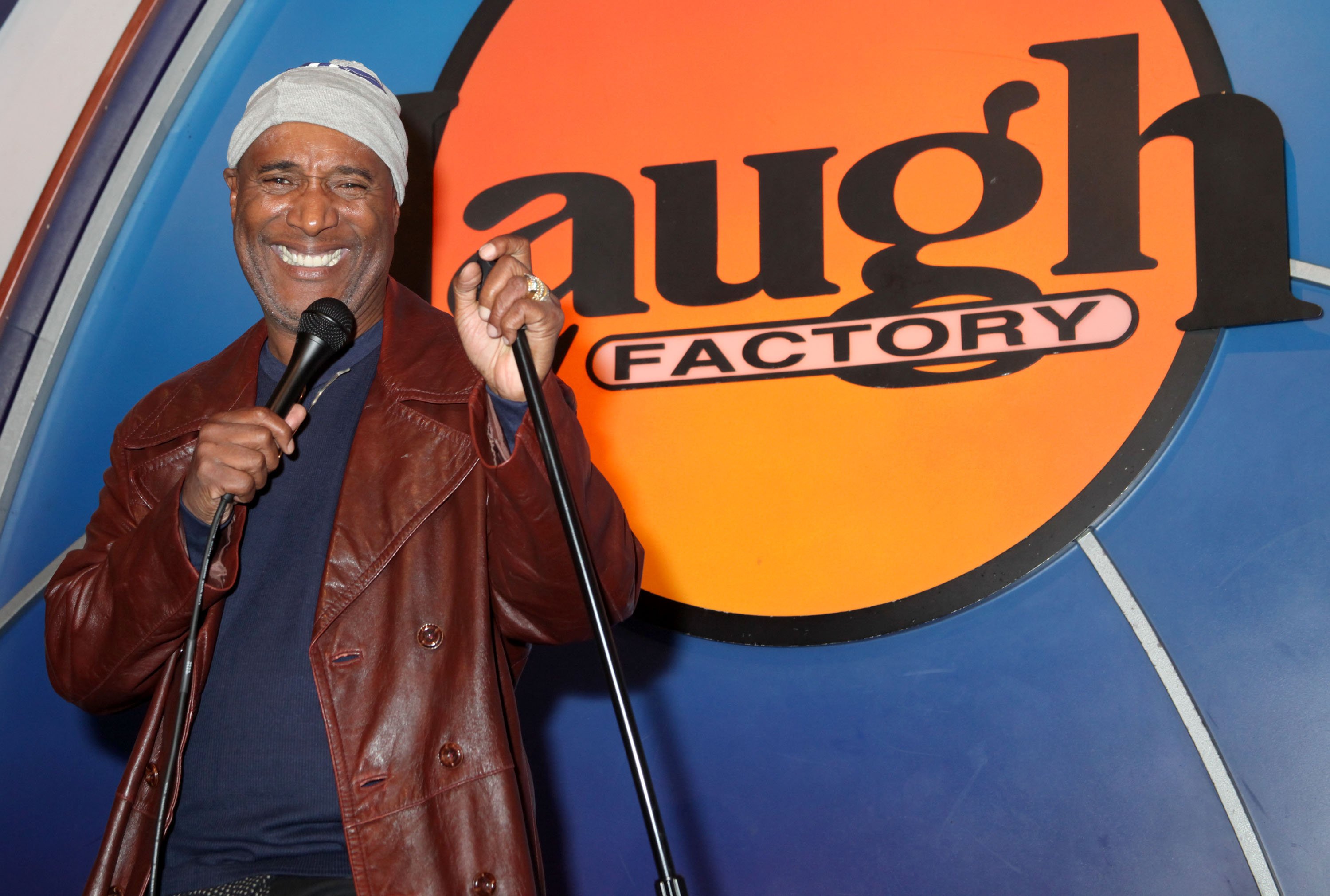 Paul Mooney in a grey beanie and brown leather jack on stage at the Laugh Factory