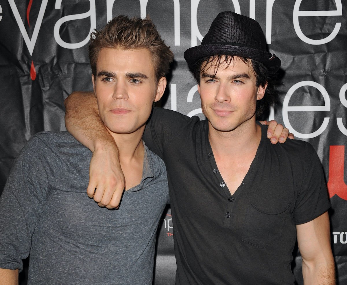 Paul Wesley and Ian Somerhalder pose at a meet and greet in grey shirts and denim jeans in front of a black backdrop that says 'The Vampire Diaries.' Somerhalder wears a fedora and is wrapping his arm around Wesley's shoulder.