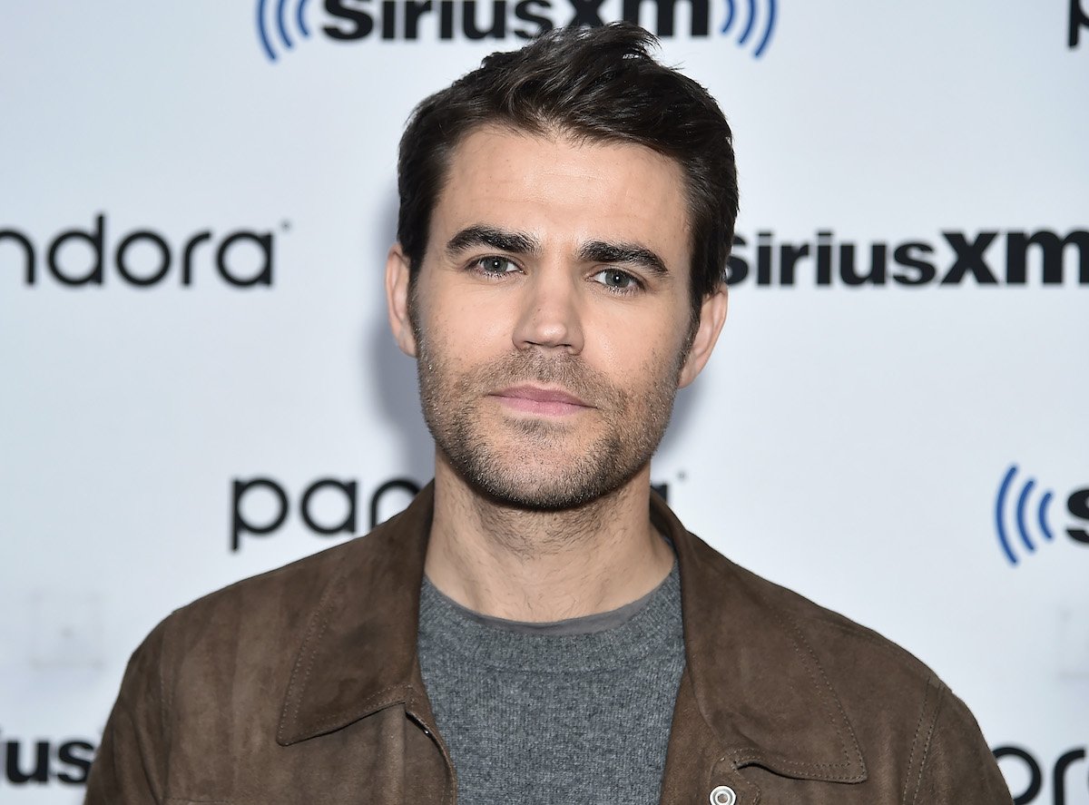 Paul Wesley in a grey shirt and brown jacket in front of a white backdrop that says 'Pandora' and 'SiriusXM Radio'