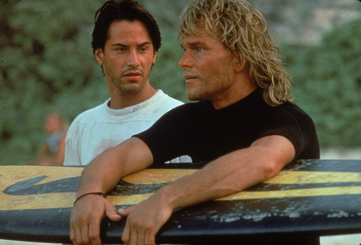 Keanu Reeves and American actor Patrick Swayze stand on a beach as Swayze holds a surfboard in 'Point Break'