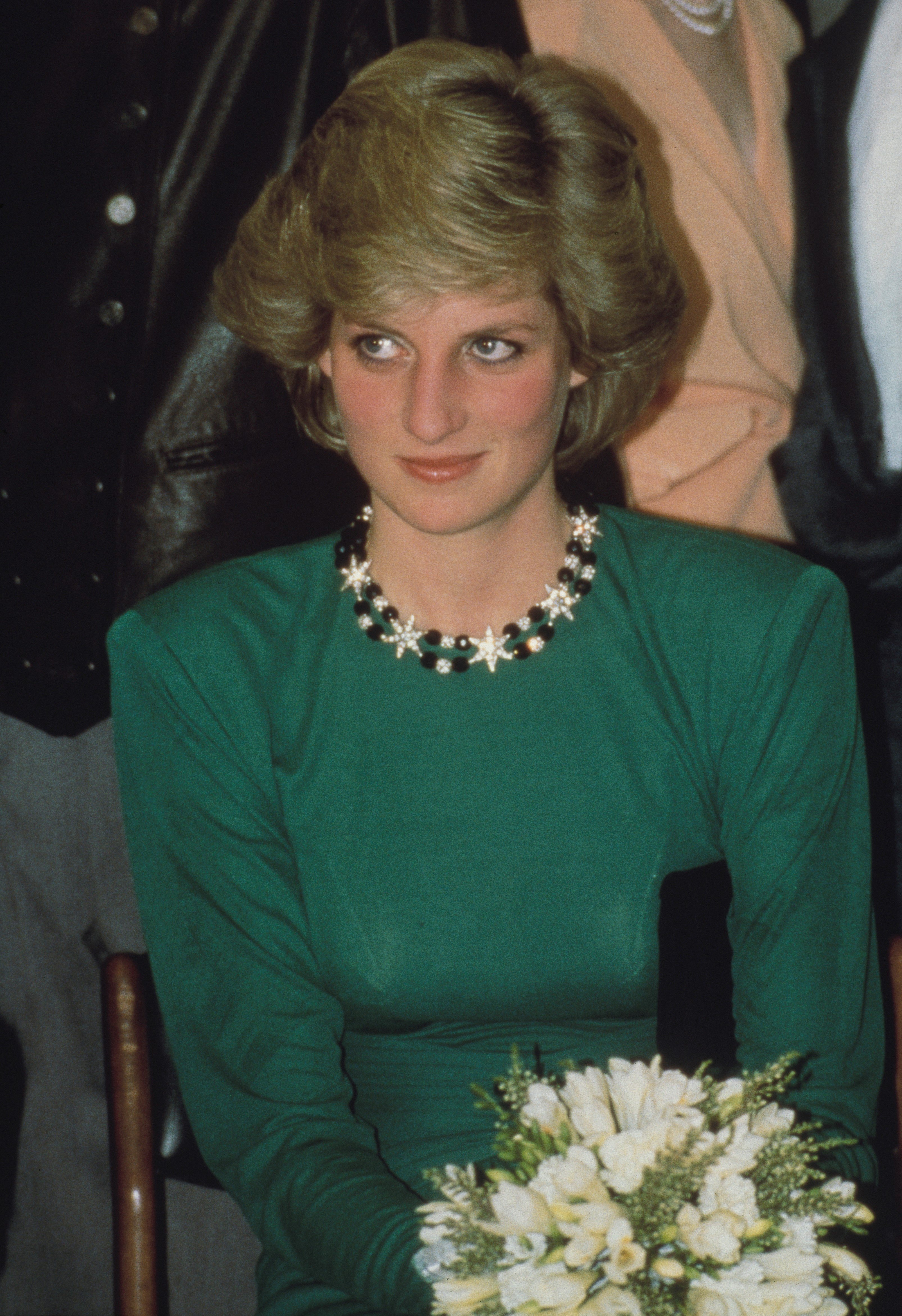 Princess Diana seated in a dark green dress and holding a bouquet of flowers at the headquarters of the London City Ballet