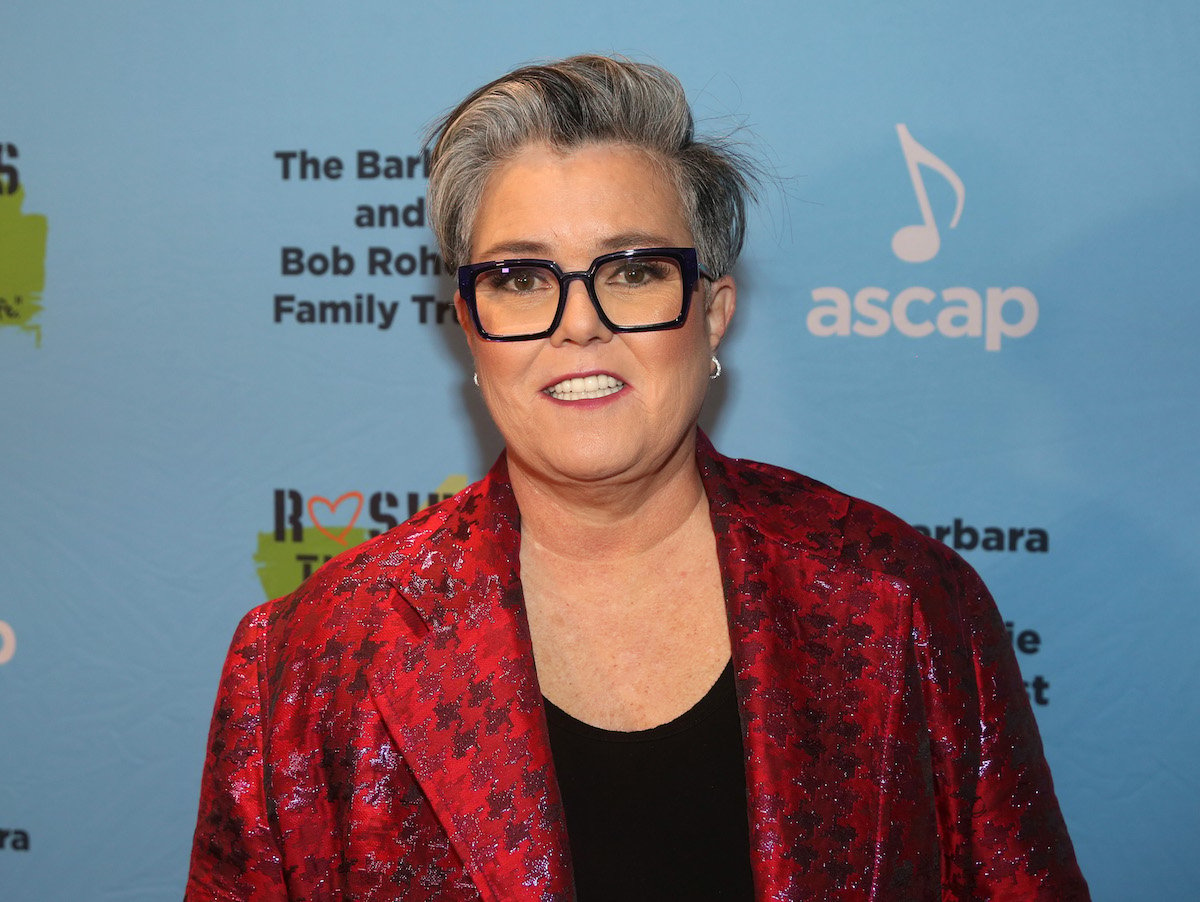 Rosie O'Donnell poses on a red carpet in a black shit and red jacket.