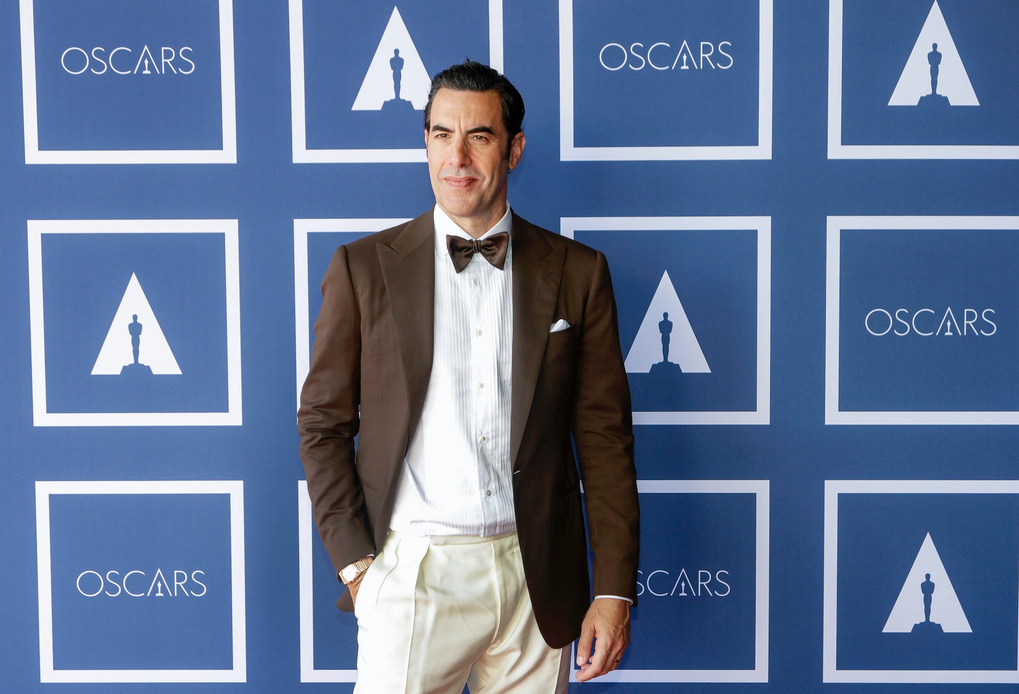 Sacha Baron Cohen smiling in front of a blue background