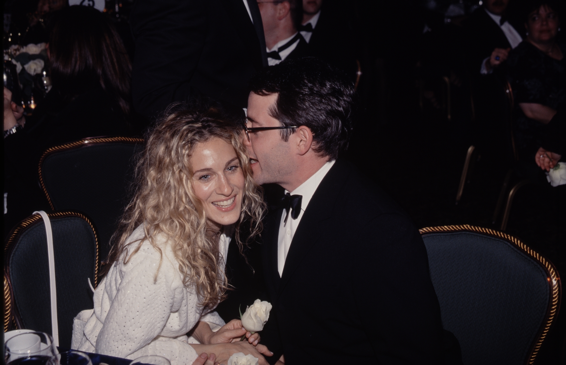 Sarah Jessica Parker smiling while her husband, Matthew Broderick, whispers in her ear at an event.