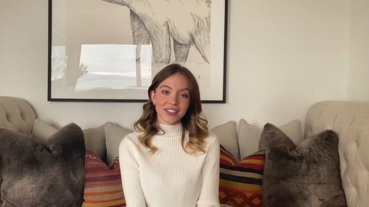 Sydney Sweeney sits on a couch in a white turtleneck