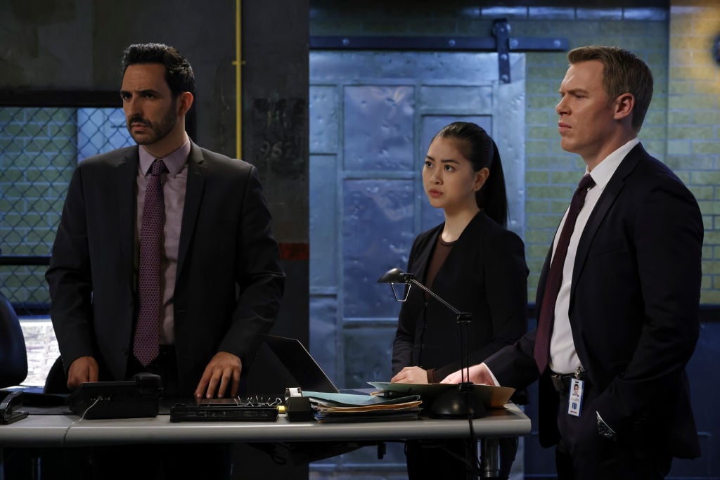 Amir Arison as Aram Mojtabai is dressed in a suit next to Laura Sohn as Agent Alina Park in all black, and Diego Klattenhoff as Donald Ressler in a suit. They all look concerned.