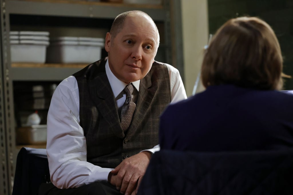 James Spader as Raymond 'Red Reddington, wearing a brown suit vest and white button-up, sits across a woman.