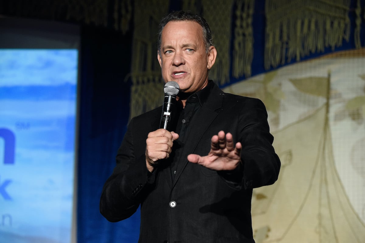 Tom Hanks holds a mic and looks serious while holding a hand out as he speaks onstage