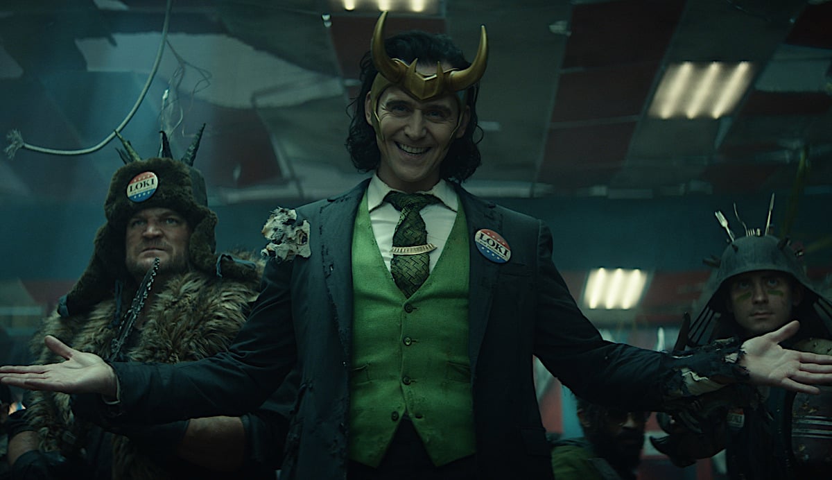 Tom Hiddleston smiles deviously in a green vest and tie, black blazer, and gold crown with a red, white, and blue pin that reads 'Loki' in Marvel's 'Loki' TV series on Disney+.