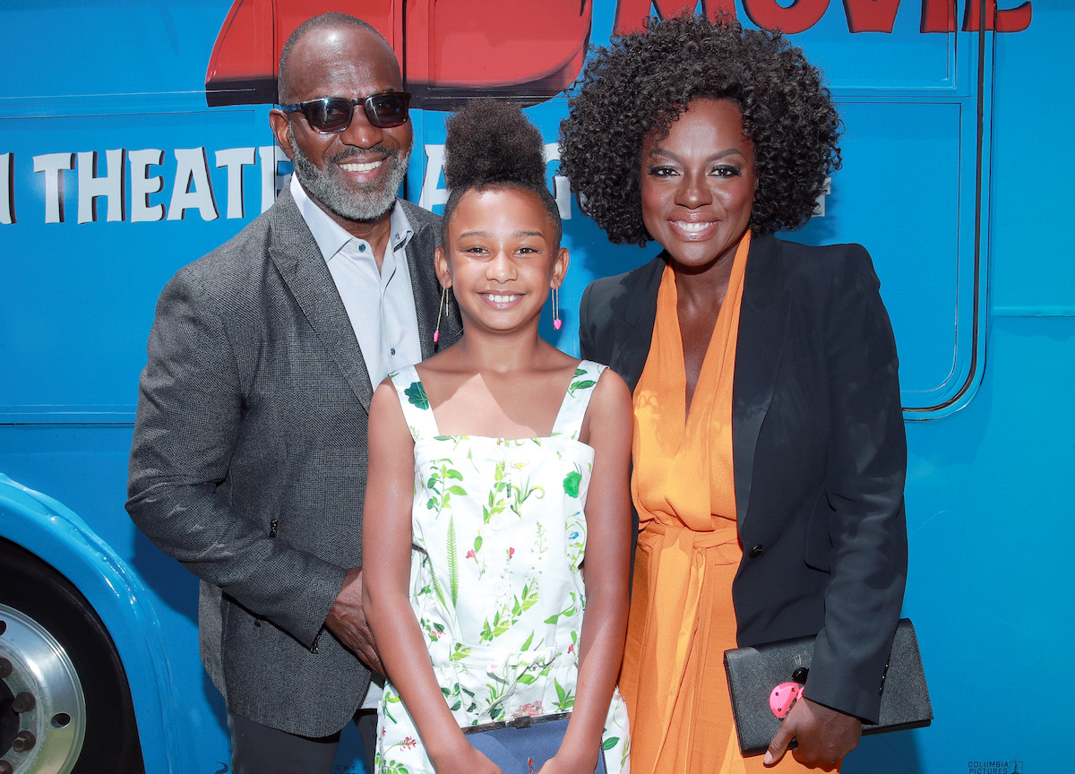 Julius Tennon smiles in sunglasses, a blue shirt, and grey suit. Genesis Tennon smiles in a white dress with a green leaf print. Viola Davis smiles in an orange dress and black blazer. They're standing in front of a blue truck that says 'The Angry Birds Movie 2''