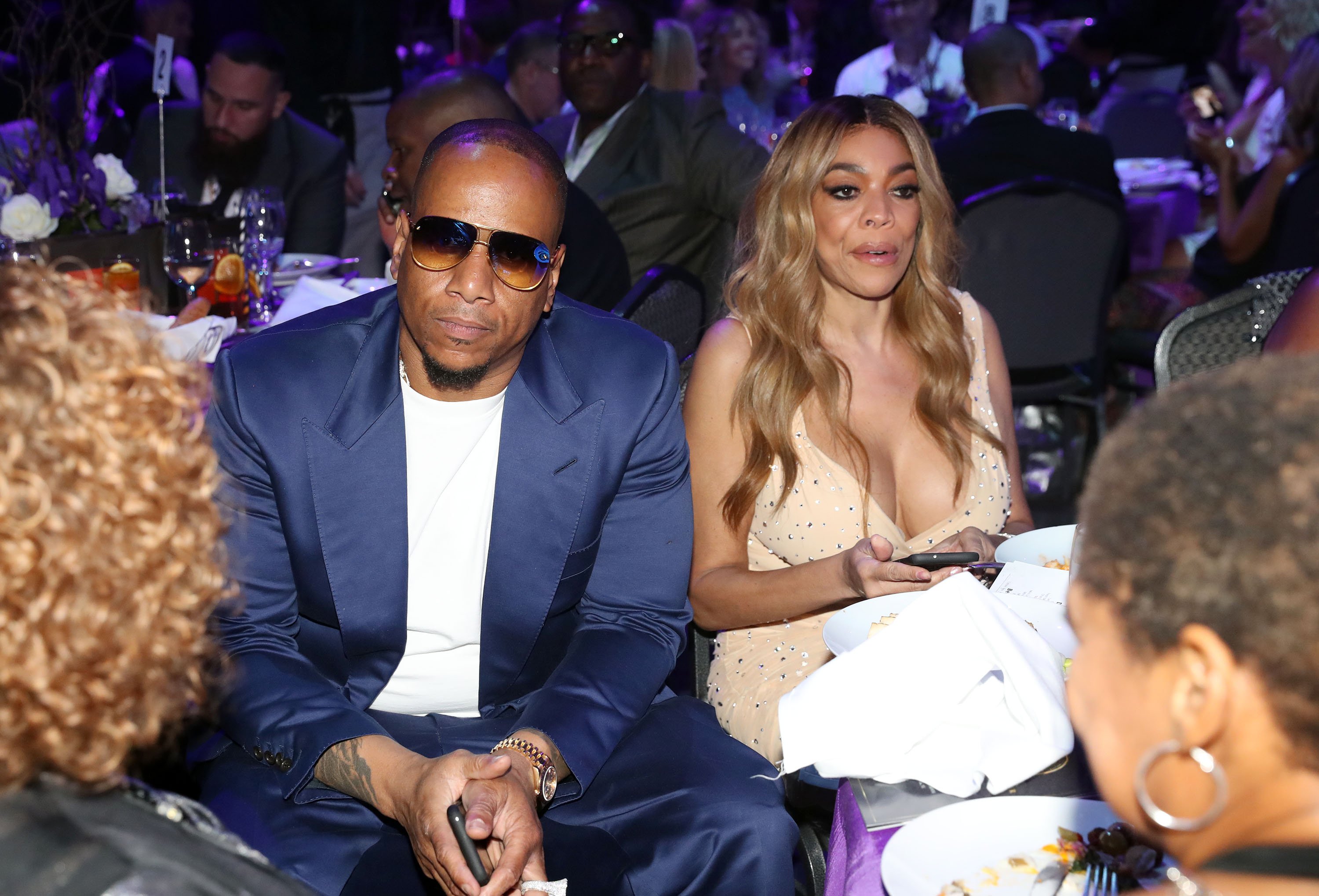 Wendy Williams looking away from the camera while wearing a gold dress and sitting next to Kevin Hunter wearing an all-blue suit.