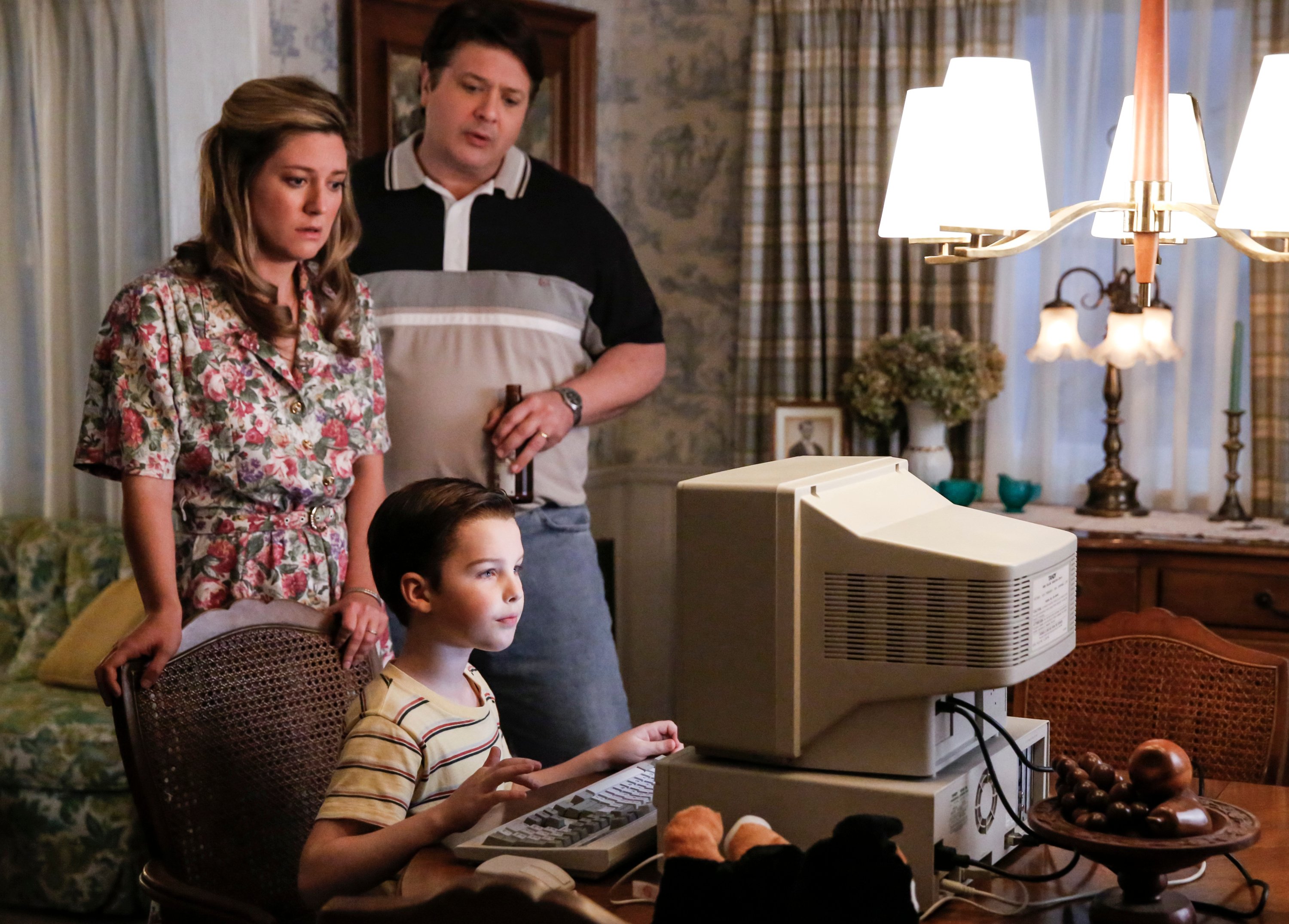 Sheldon Cooper sits at a computer in the Cooper family home. Mary and George Cooper stand behind him in an episode of 'Young Sheldon'