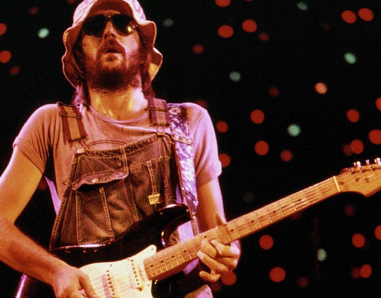 Eric Clapton plays guitar wearing overalls and sunglasses on stage in 1974