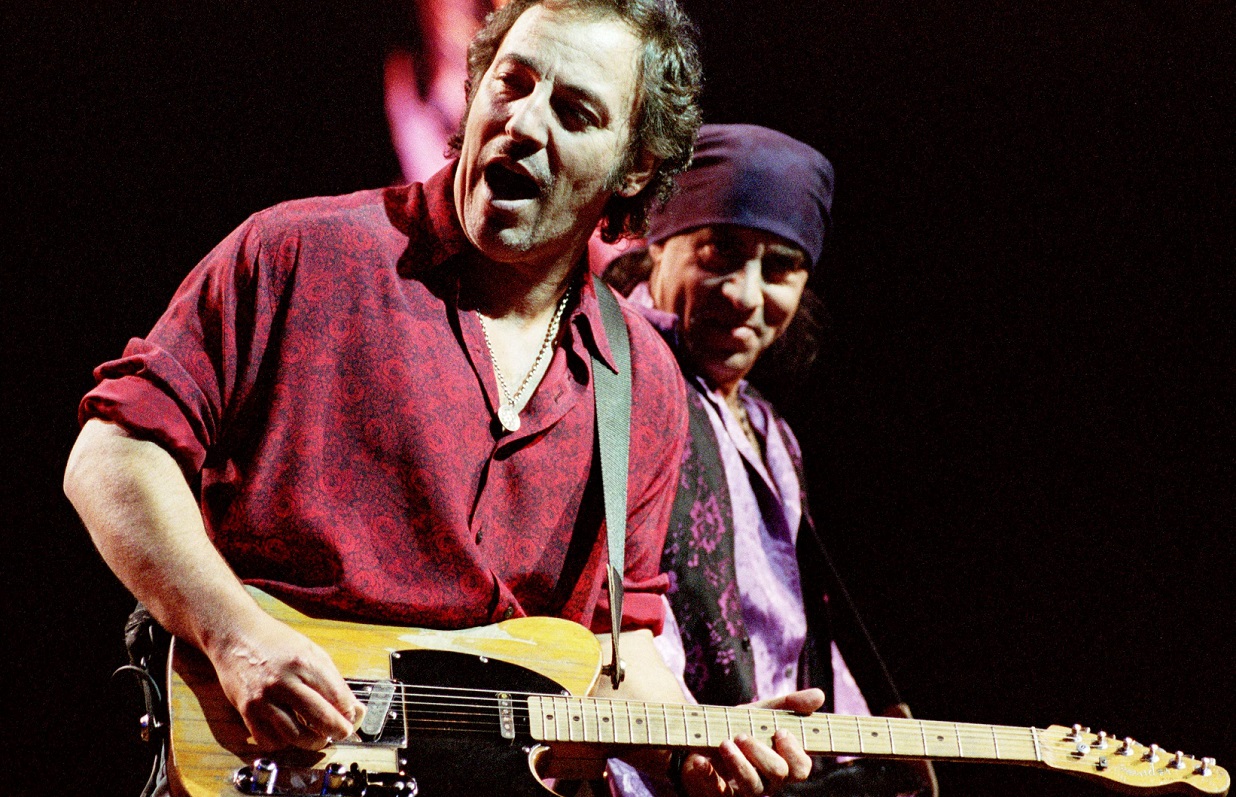 Bruce Springsteen sings and plays guitar on stage as Steven Van Zandt plays guitar and smiles at Springsteen.