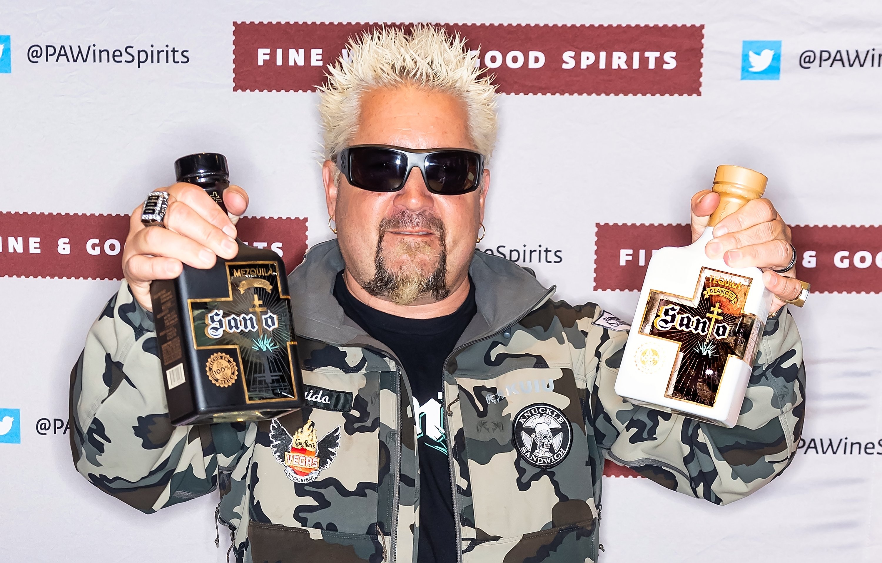 Guy Fieri, wearing sunglasses, poses with bottles of his Santo Tequila