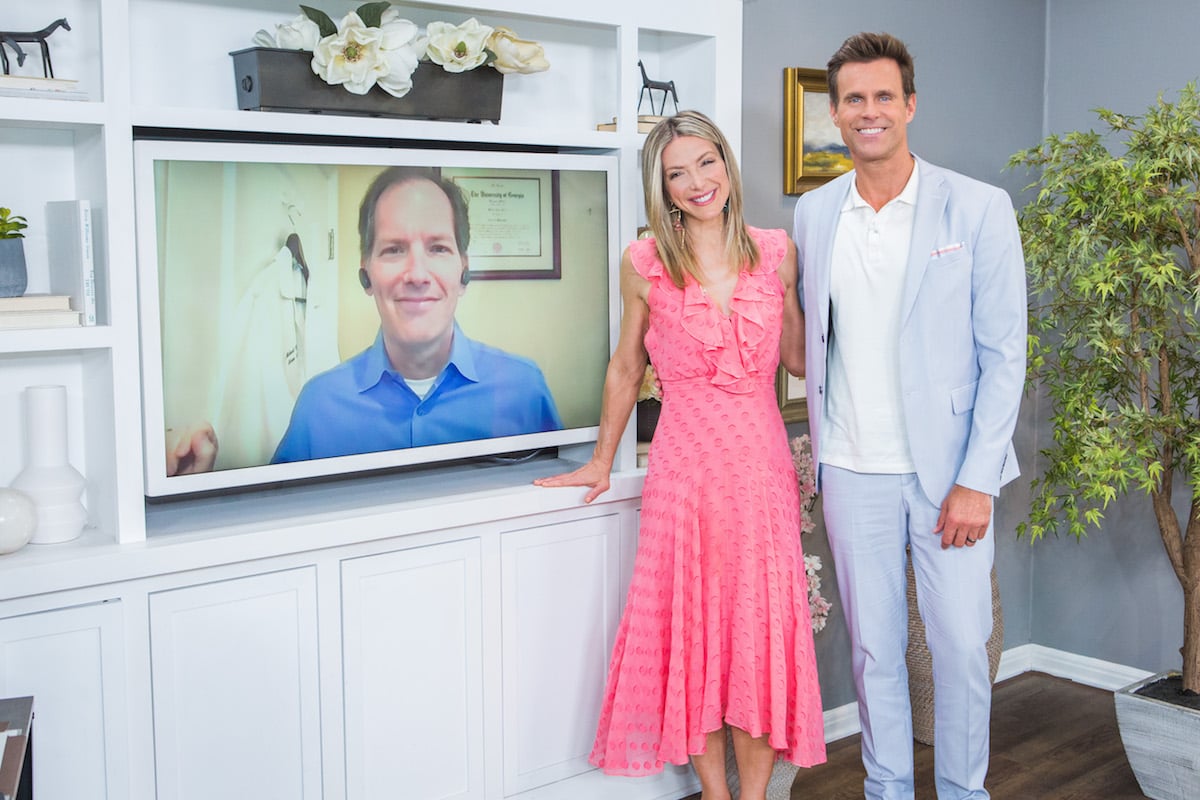 Home and Family hosts Debbie Matenopoulos and Cameron Mathison standing next to a TV screen 