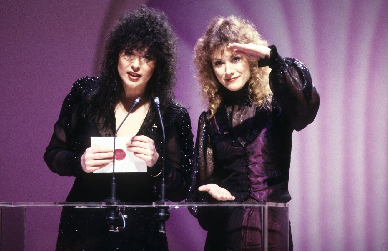 Ann Wilson holds an envelope and speaks into the microphone as Nancy Wilson gestures next to her at a podium.