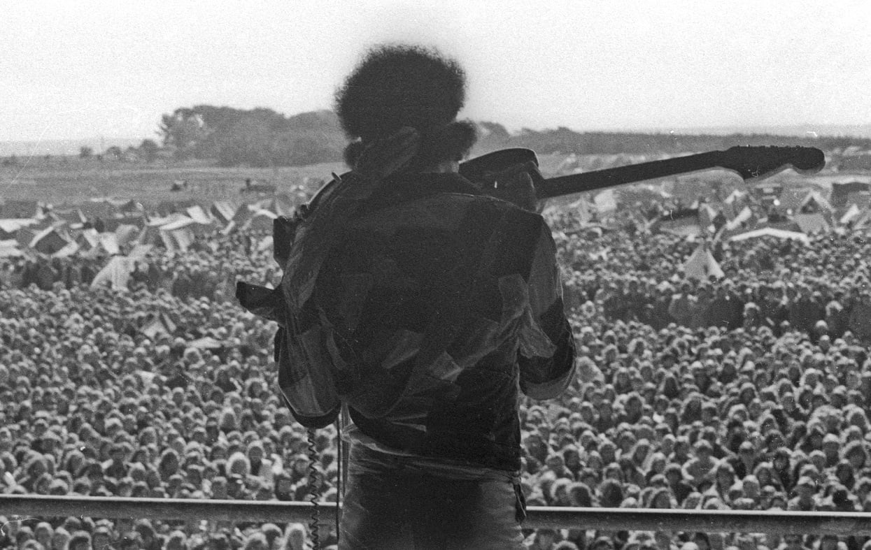 View from behind of Jimi Hendrix playing the guitar with his teeth, with a sea of fans in the background