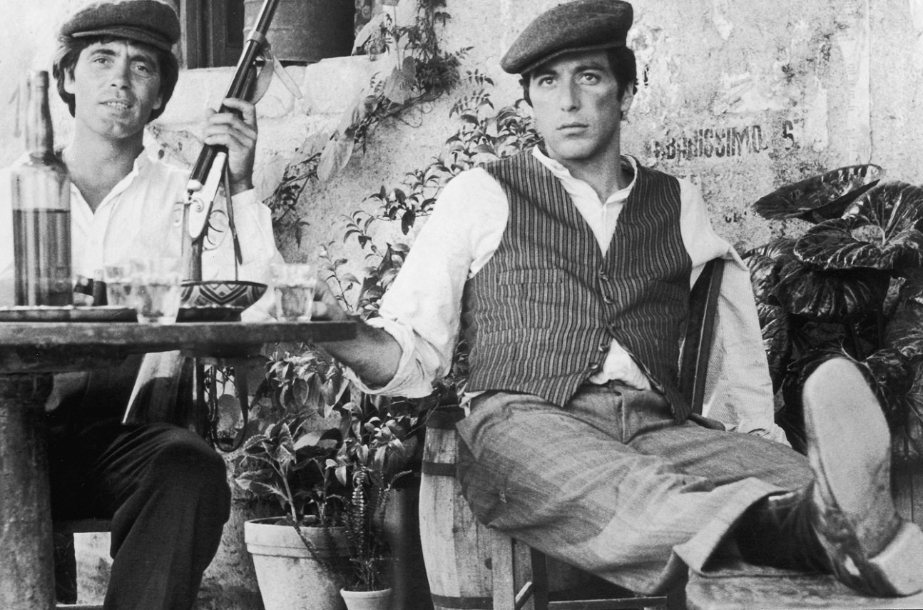 Al Pacino sits with Franco Citti, who holds a rifle at an outdoor table in 'The Godfather'. Pacino rests his leg on a chair. 