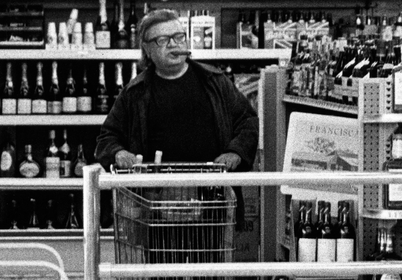 Mario Puzo, with cigar in his mouth, pushes a shopping cart through the aisles of a wine store.