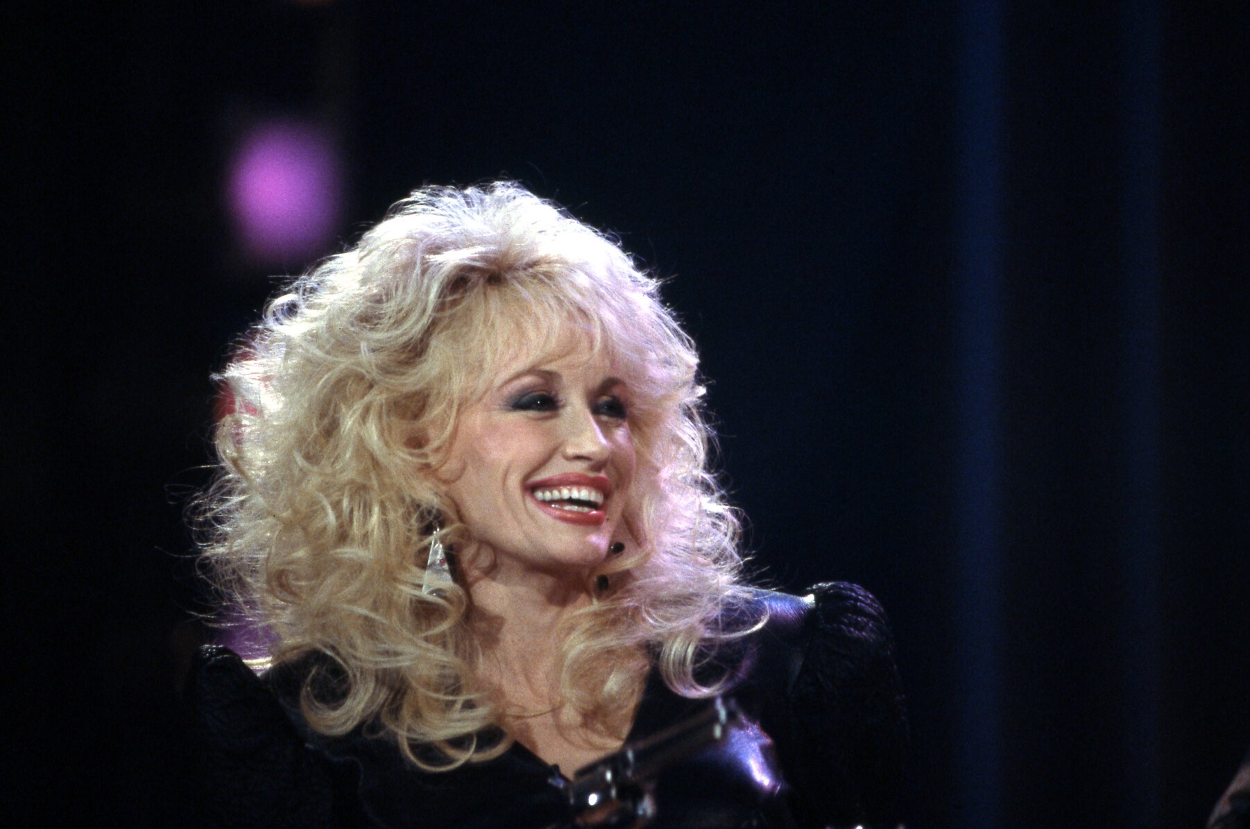 Dolly Parton in 1988. She's wearing a black blouse, big, teased hair, and an open-mouthed smile on her face. She's in front of a black background.