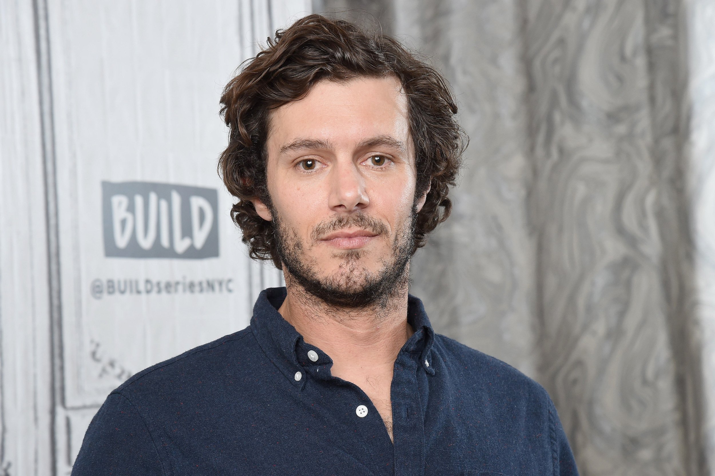 Adam Brody wears a dark blue shirt and stands in front of a Build Series wall
