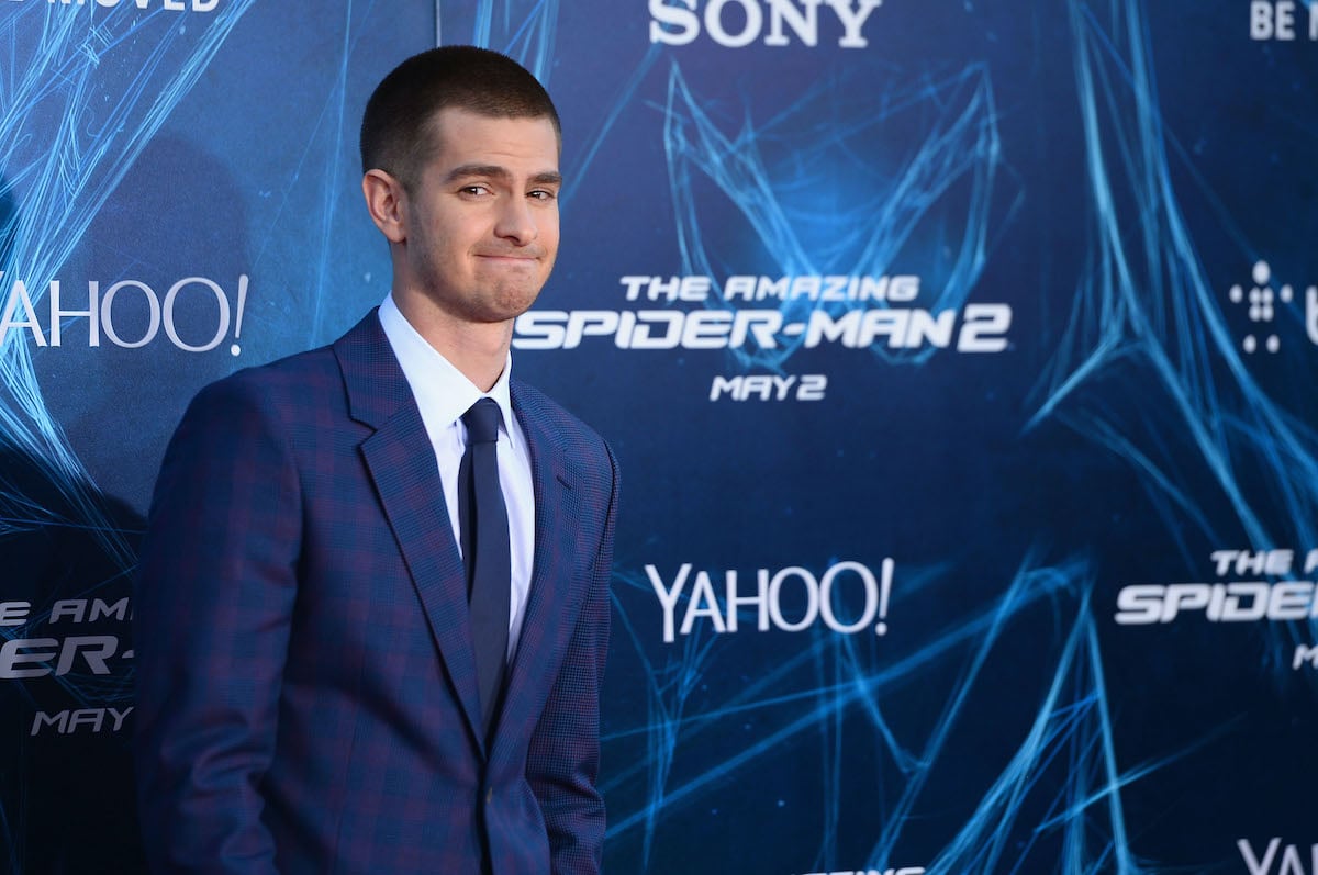 Andrew Garfield wears a blue suit and stands in front of a logo display of 'The Amazing Spider-Man 2'