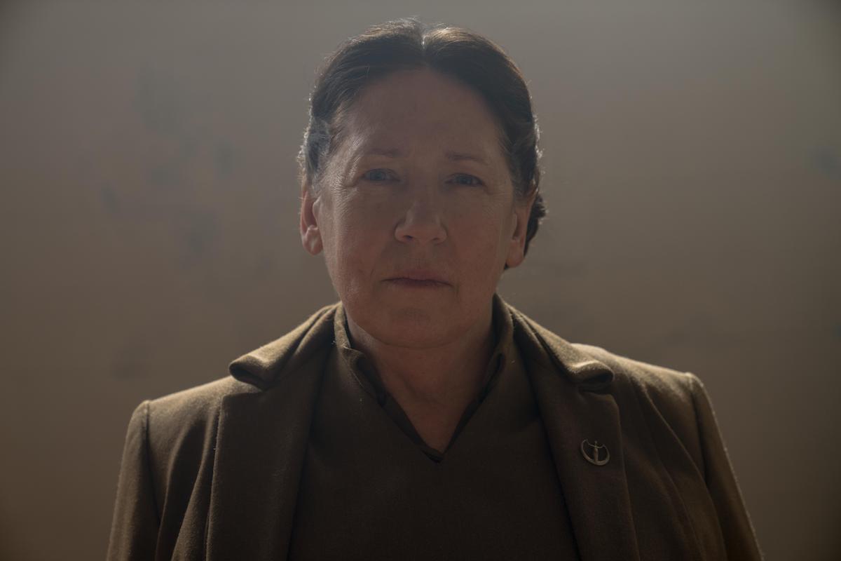 Ann Dowd as Aunt Lydia in 'The Handmaid's Tale' Season 3. She looks directly at the camera with a powerful look on her face. She wears a brown dress and brown overcoat with a gold Gilead pin on her lapel. Her hair is pulled back in a bun. She stands in front of a beige wall.