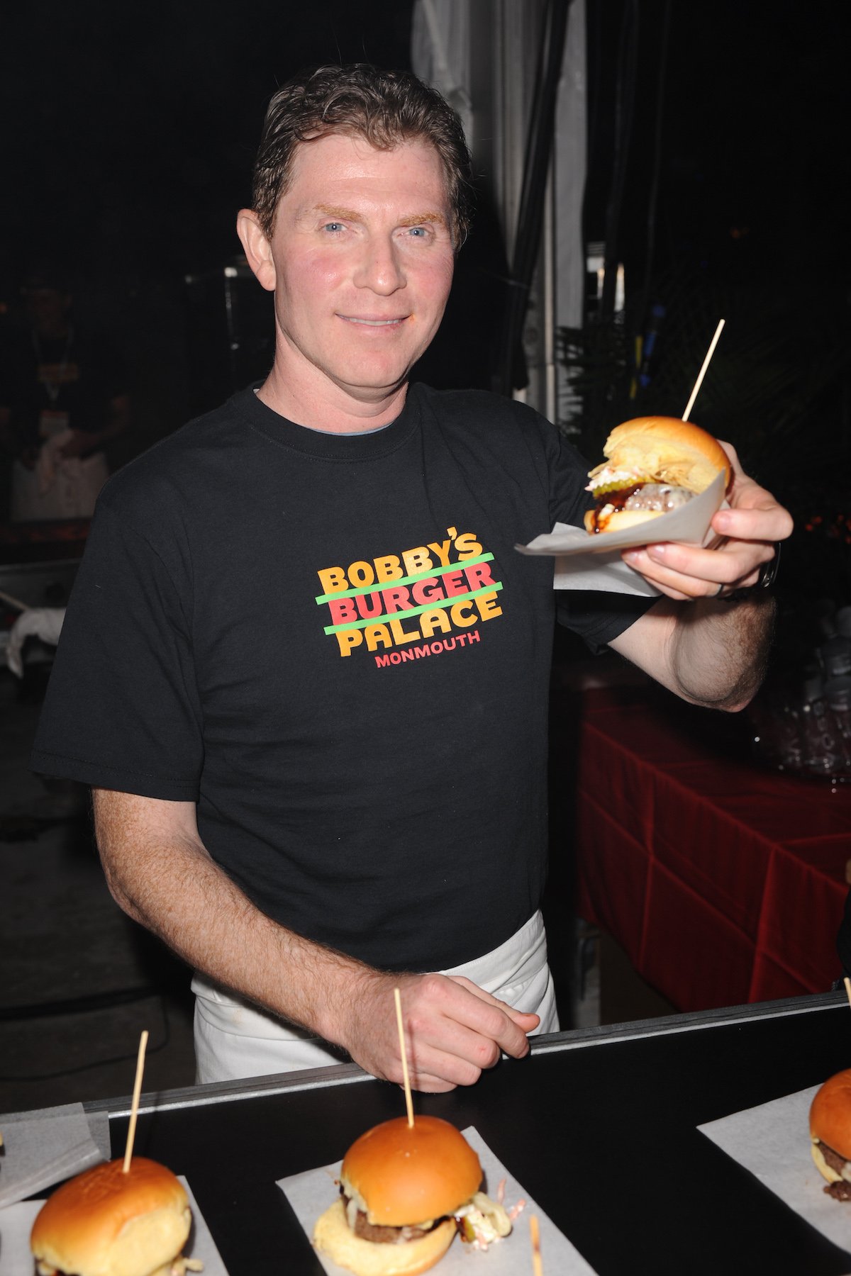 Bobby Flay smiles as he holds up a hamburger