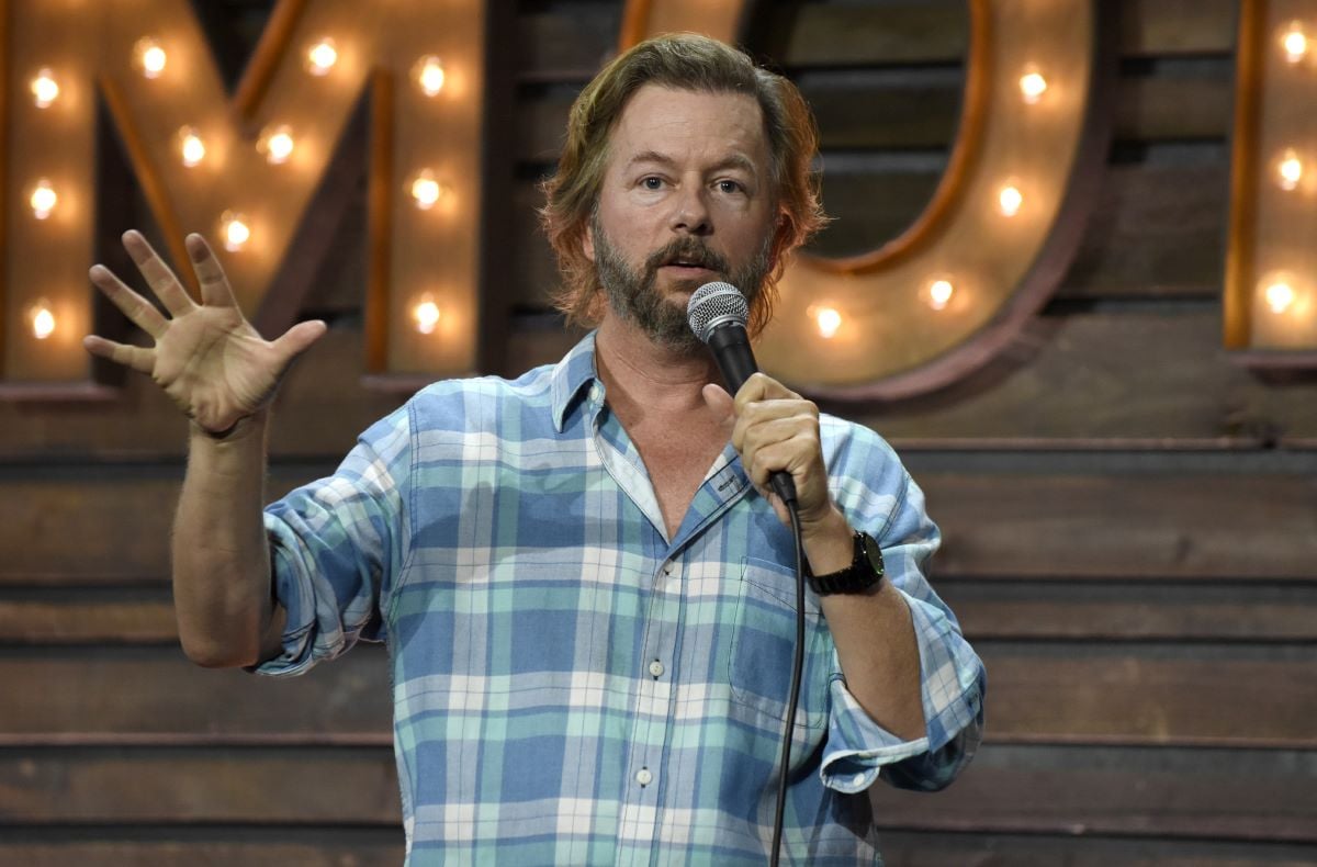 New 'Bachelor in Paradise' host David Spade performs with a microphone while wearing a plaid blue shirt